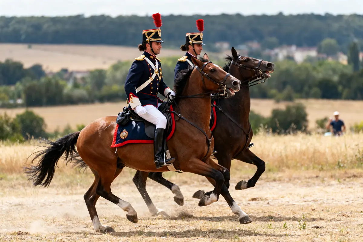 Deux cavaliers en uniforme historique chevauchent à travers champ, portant des ornements rouges et bleus.