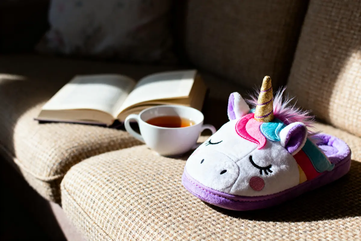 Chausson licorne coloré sur canapé éclairé, tasse de thé et livre ouvert en arrière-plan.